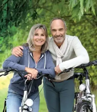 a middle aged couple are leaning on their bicycles while out for a ride on a forested trail.