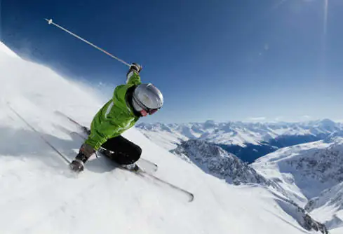 a skier wearing a helmet and a green ski jacket is seen skiing down a mountain with a wide vista of the mountain range in the background of a blue sky
